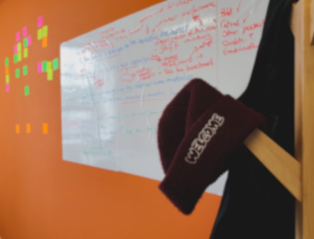 A maroon hat is placed on a coat rack with a whiteboard and some stickynotes in the background on an orange wall