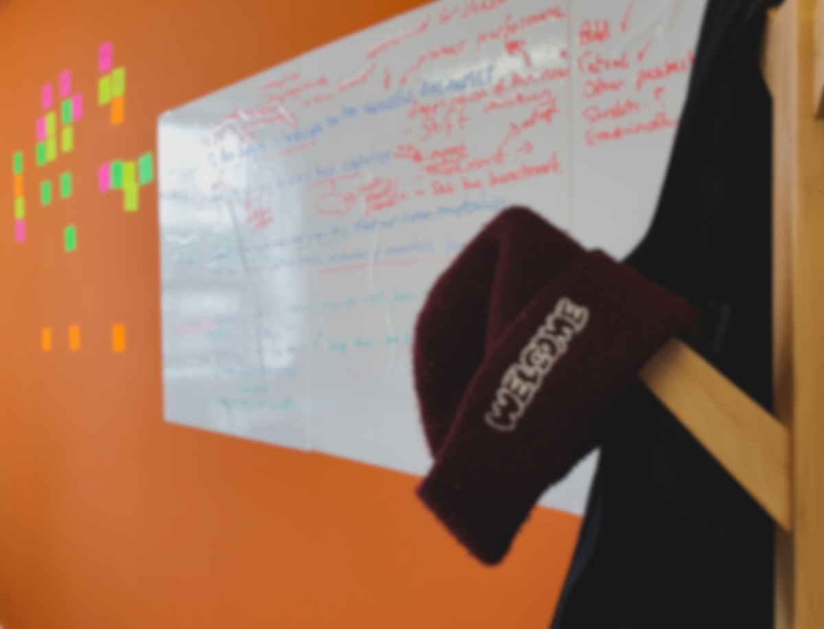 A maroon hat is placed on a coat rack with a whiteboard and some stickynotes in the background on an orange wall